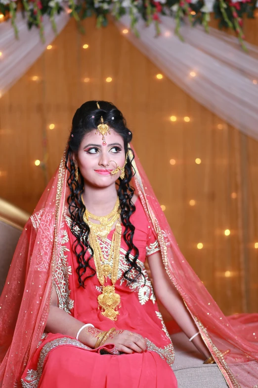 a woman in a red dress sitting on a couch, hurufiyya, wearing a wedding dress, long braided hair on top of head, wearing gold jewellery, with professional lighting