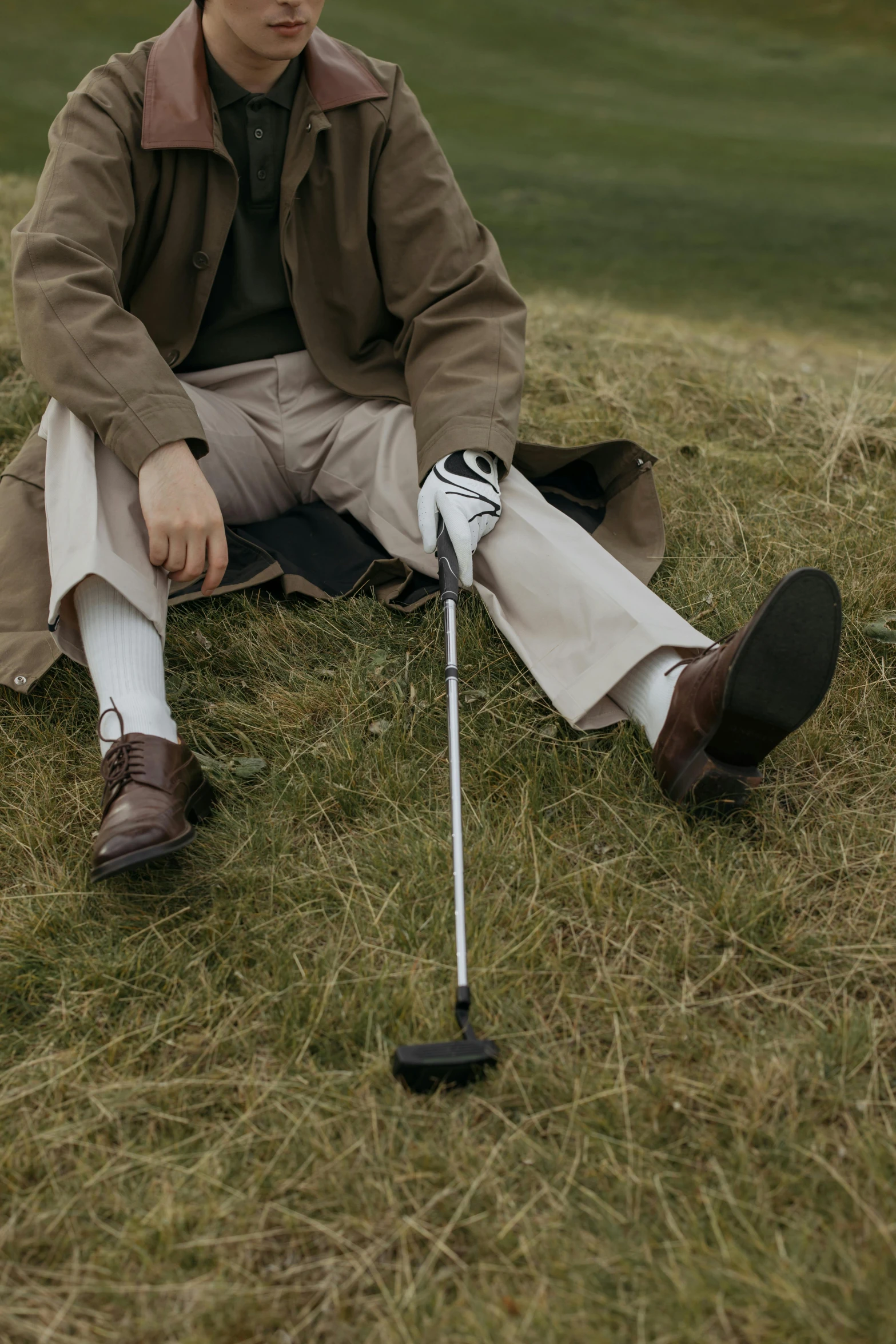 a man sitting on top of a grass covered field, holding a cane, gray shorts and black socks, muted brown, thumbnail