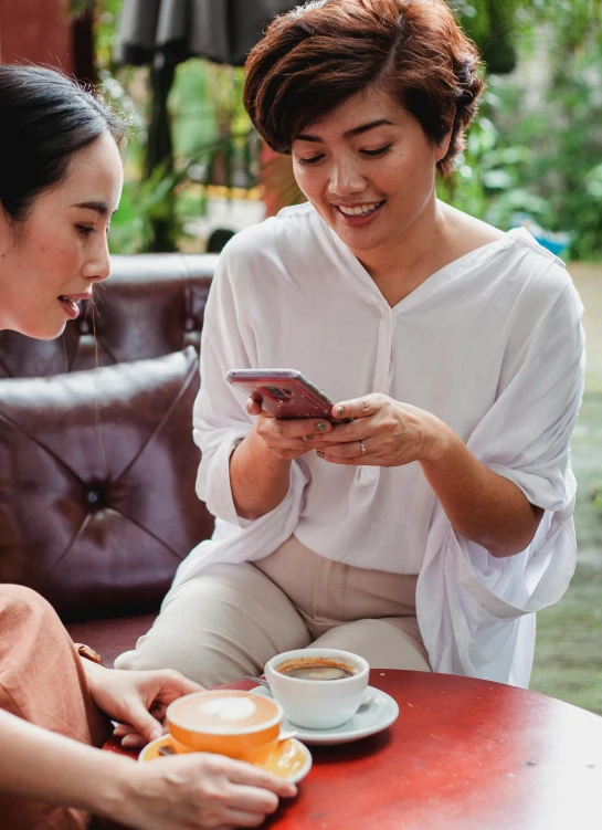 two women sitting at a table looking at a cell phone, trending on pexels, indonesia, square, sitting on a mocha-colored table, avatar image