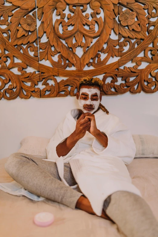 a man sitting on a bed with a mask on, by Arabella Rankin, trending on pexels, deep tan skin, white facepaint, spa, having a great time