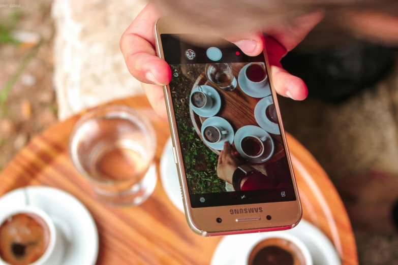 a person taking a picture of a cup of coffee, by Julia Pishtar, pexels contest winner, android phones, avatar image, sitting on a mocha-colored table, gold