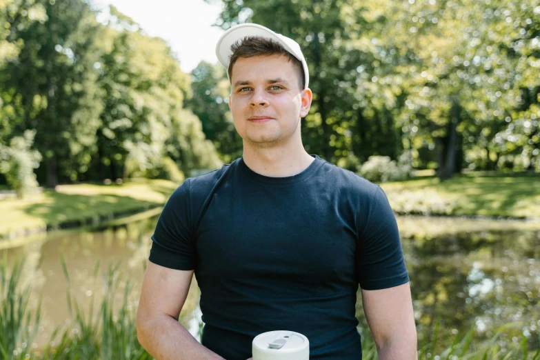 a man standing in front of a pond holding a cup of coffee, a portrait, by Adam Marczyński, he is wearing a black t-shirt, avatar image, professional profile photo, he is about 20 years old | short