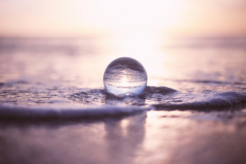 a glass ball sitting on top of a sandy beach, purple water, blurred, sun rising, inside a marble