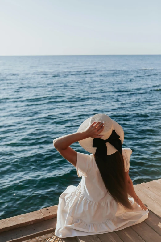 a woman sitting on a dock looking out at the ocean, pexels contest winner, wearing hair bow, white hat, wearing white silk, twirling