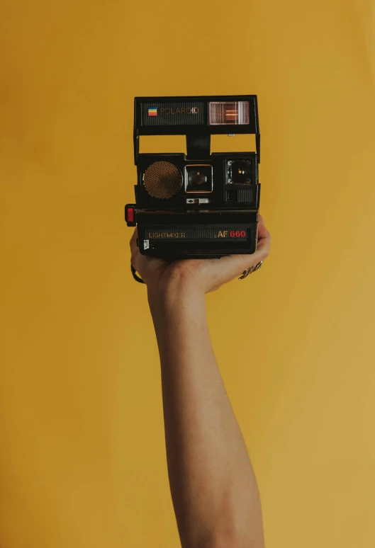 a person holding a polaroid camera in their hand, inspired by Elsa Bleda, unsplash contest winner, postminimalism, black and yellow and red scheme, holding gold, on a yellow canva, instagram post