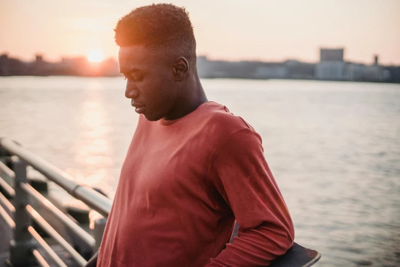 a man standing next to a body of water holding a skateboard, pexels contest winner, happening, handsome hip hop young black man, looking off into the sunset, with red haze, slightly muscular