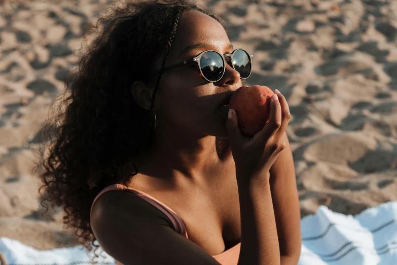 a woman eating an apple on the beach, a portrait, trending on pexels, brown skinned, wearing shades, 1 2 9 7, low light