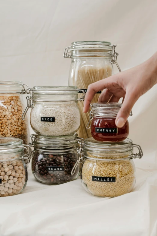 a table topped with jars filled with different types of food, by Nina Hamnett, pexels, cereal, label, plain background, sleek hands