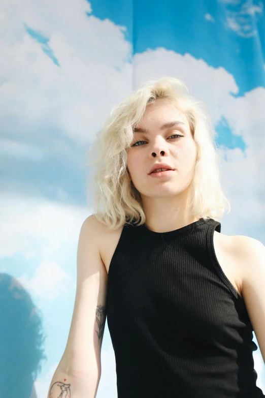 a woman with blonde hair standing in front of a painting, an album cover, inspired by Elsa Bleda, unsplash, she is wearing a black tank top, portrait anya taylor-joy, with clouds in the sky, short platinum hair tomboy