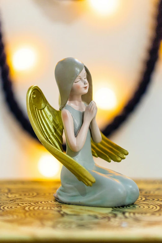 a figurine of an angel sitting on a table, golden aura, with celadon glaze, prayer hands, night setting
