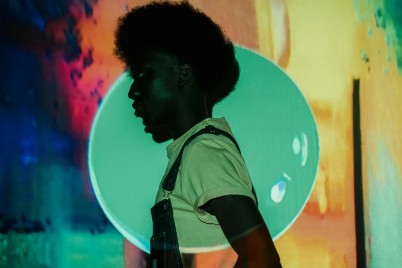 a man standing in front of a colorful wall, an album cover, inspired by Gordon Parks, pexels contest winner, funk art, large diffused glowing aura, black man with afro hair, infinity mirror, artist wearing overalls
