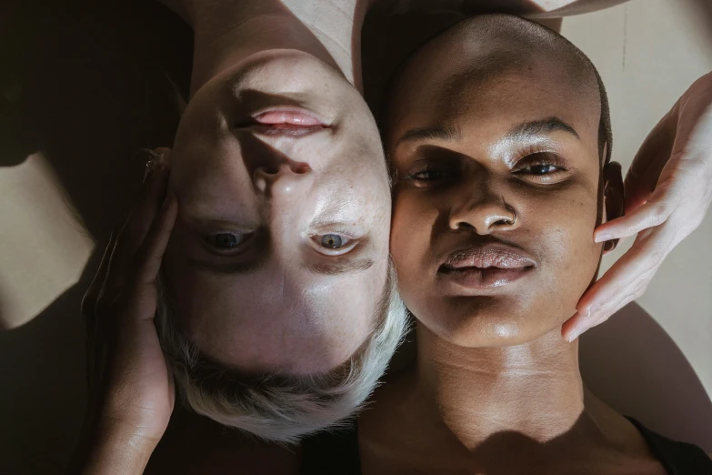 a couple of women laying on top of each other, a portrait, by Matija Jama, trending on pexels, hyperrealism, shaven face, varying ethnicities, reflective skin, looking up at the camera