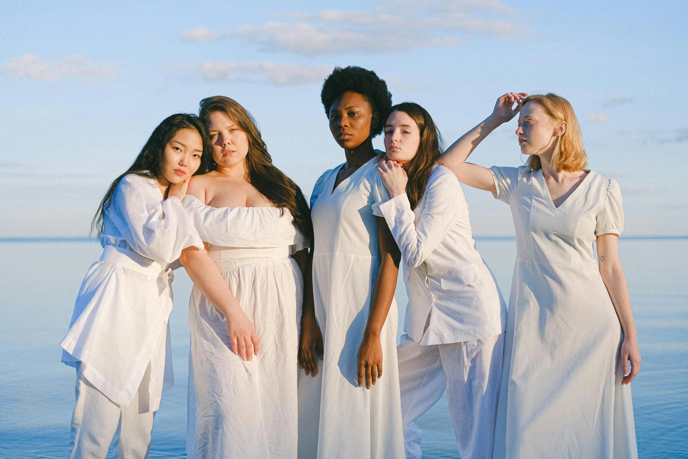 a group of women standing next to each other on a beach, an album cover, unsplash, renaissance, white porcelain skin, white uniform, varying ethnicities, embracing