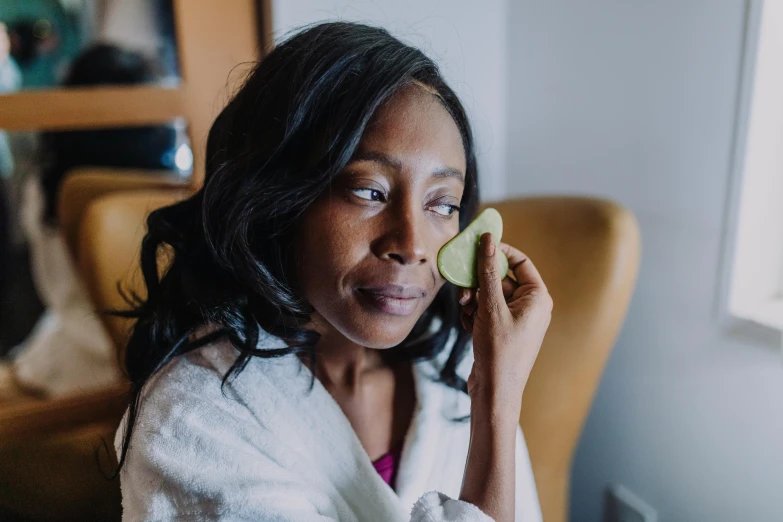 a woman holding a cucumber up to her face, someone sits in bed, dark skinned, wearing a simple robe, profile image