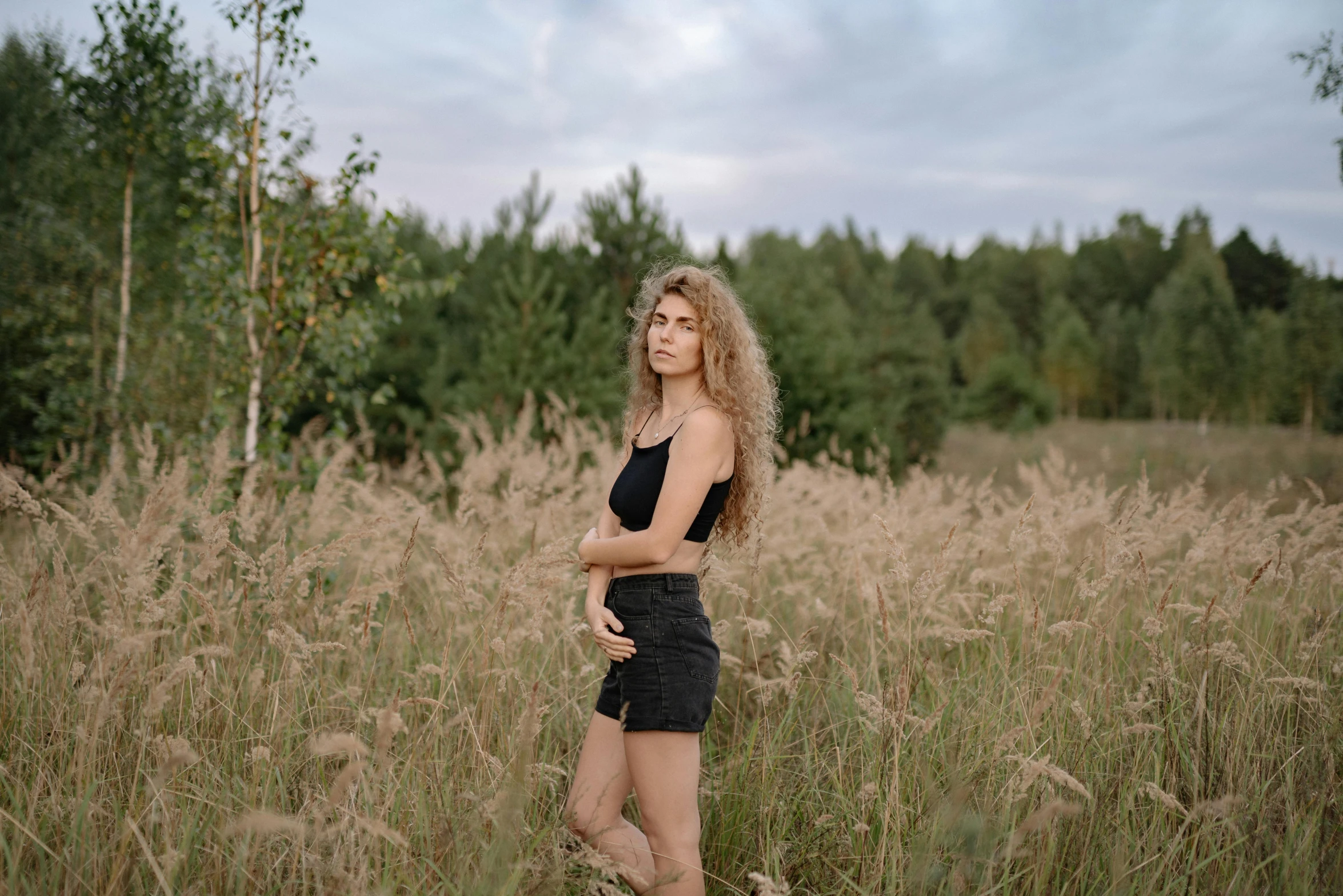 a woman standing in a field of tall grass, by Emma Andijewska, pexels contest winner, tight black tank top and shorts, russian girlfriend, curly haired, postprocessed)