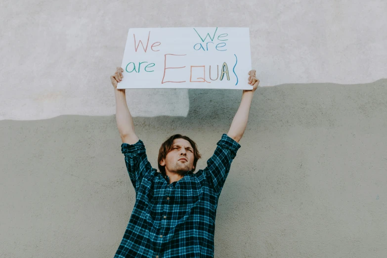 a man holding up a sign in front of a wall, by Julia Pishtar, pexels, balancing the equation, queer, sebastian stan, square