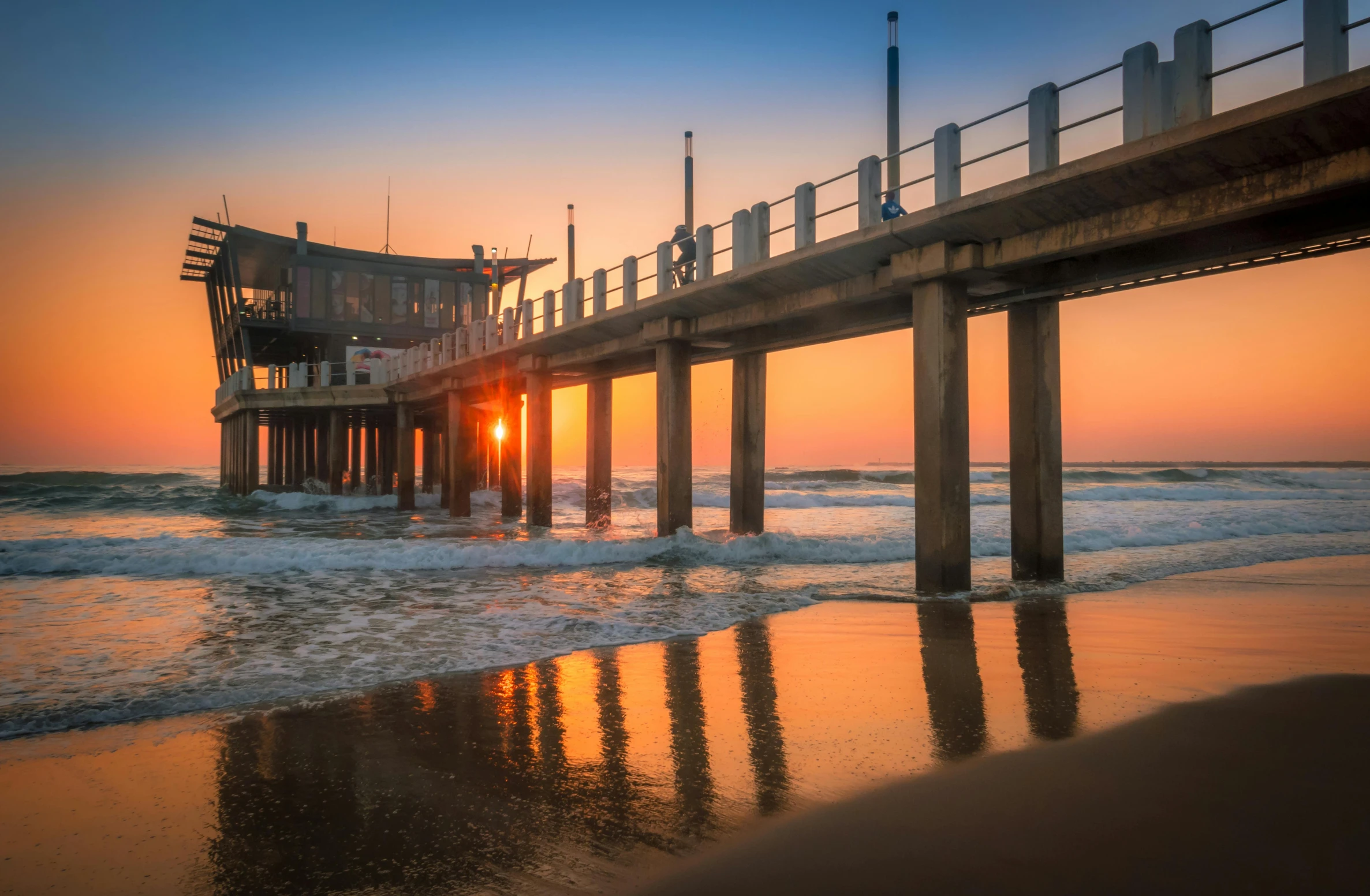 a pier sitting on top of a beach next to the ocean, by Niko Henrichon, pexels contest winner, renaissance, water reflecting suns light, south african coast, 8k hdr sunset lit, urban surroundings