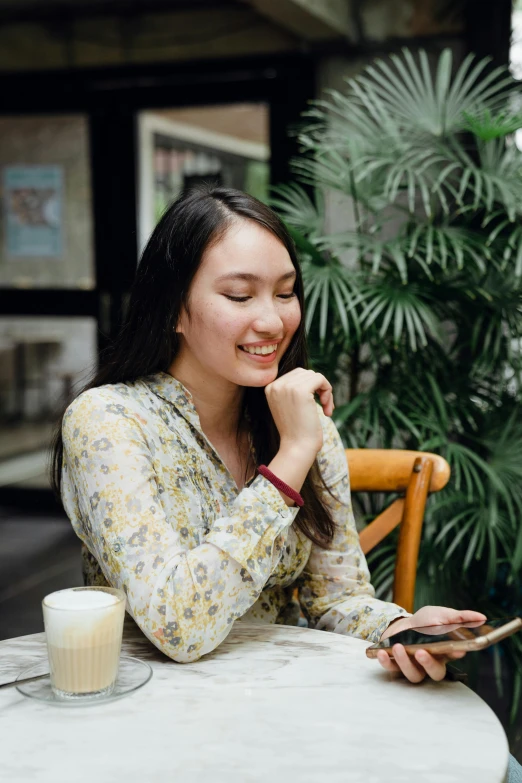 a woman sitting at a table using a cell phone, wearing yellow floral blouse, jakarta, smiling slightly, high-quality photo