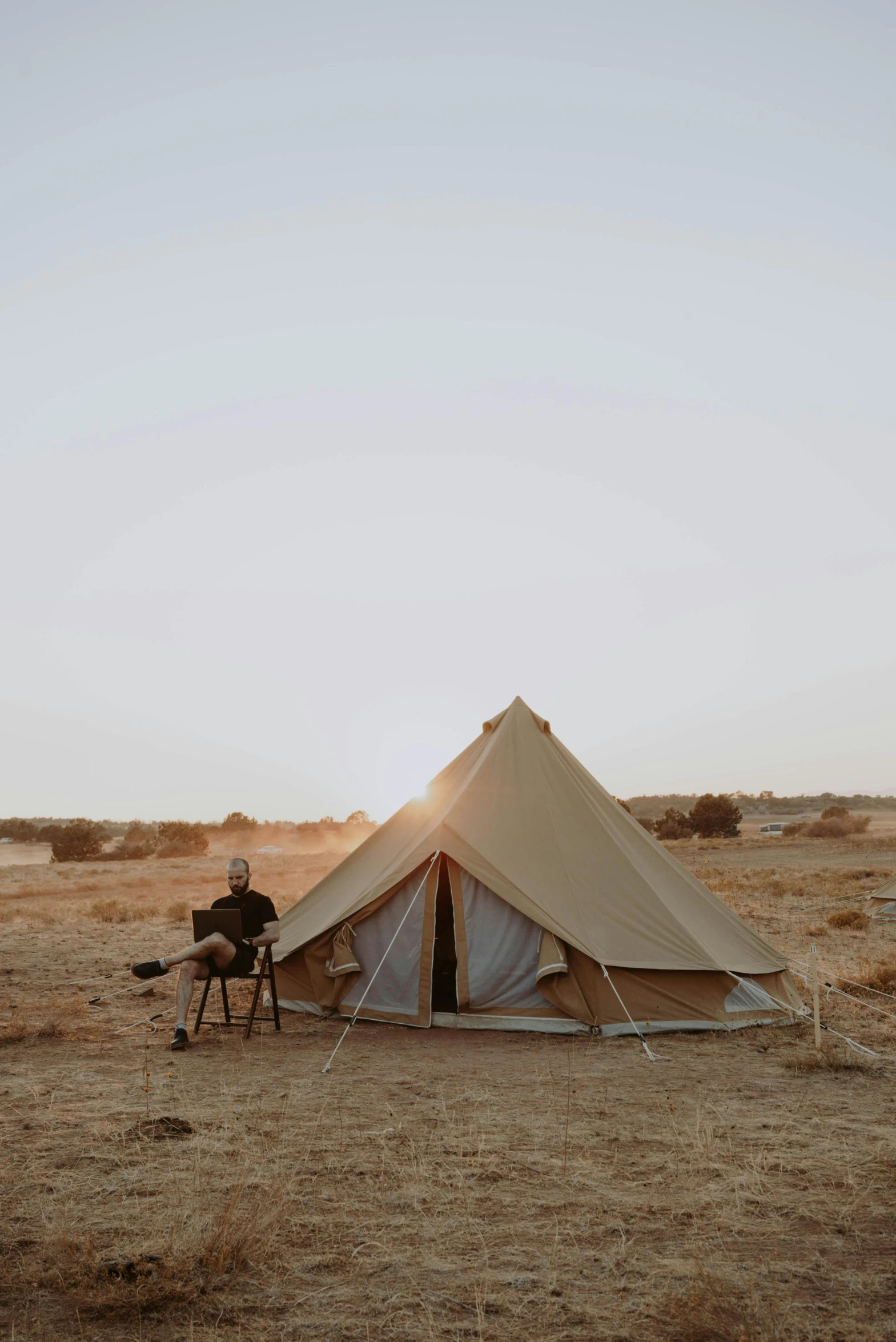 a man sitting in a chair in front of a tent, by Jessie Algie, unsplash contest winner, in the australian desert, light tan, tent architecture, 1 6 x 1 6