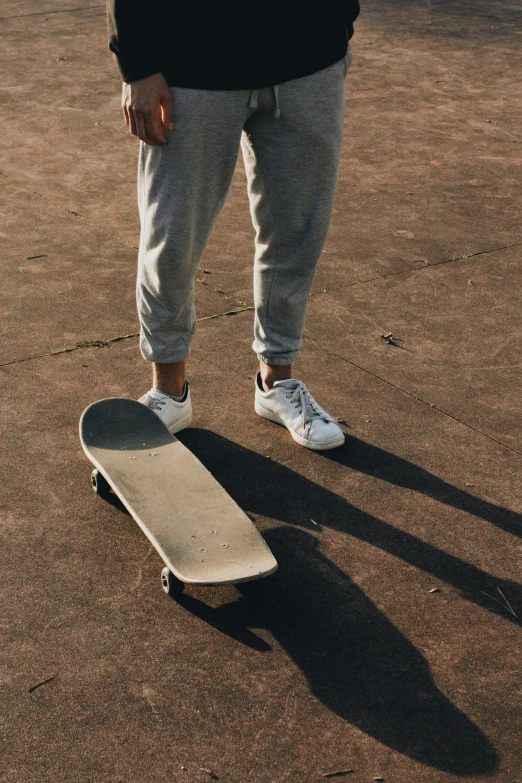 a man standing next to a skateboard on a dirt ground, trending on unsplash, white pants, solid grey, plain background, at a skate park