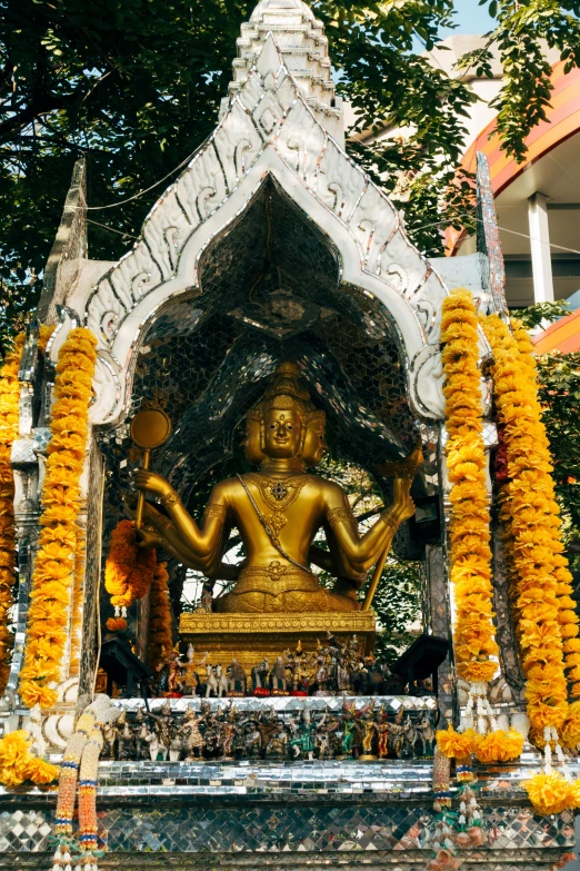 a statue of a person sitting in front of a building, a statue, samikshavad, gold flowers, elaborate cult robes, with yellow flowers around it, archways made of lush greenery