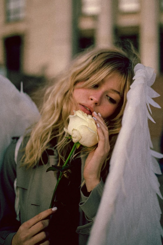 a woman holding a white rose in front of a building, inspired by Andrew Boog Faithfull, tumblr, girl with angel wings, kate moss, imogen poots as holy paladin, festivals