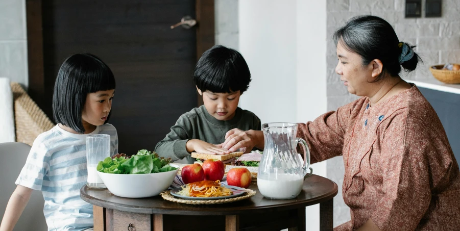 a woman and two children sitting at a table with food, pexels contest winner, asian descent, background image, kids playing, standing still