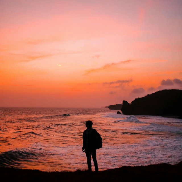 a man standing on top of a beach next to the ocean, pexels contest winner, sunset red and orange, azores, profile image, pink sunset hue