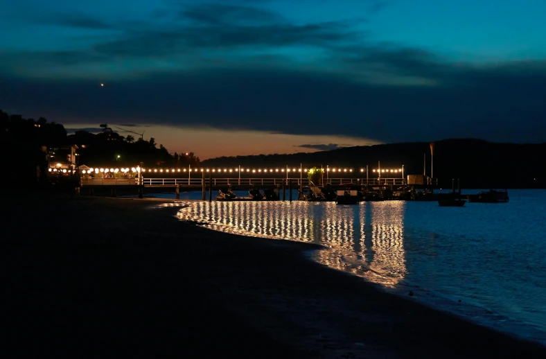 a beach at night with lights reflecting in the water, boat dock, manly, string lights, dim dusk lighting