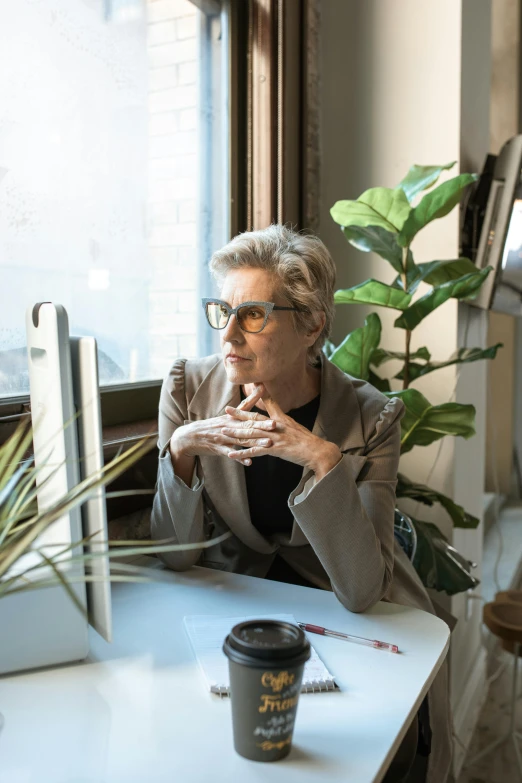 a woman sitting at a desk in front of a computer, by Richmond Barthé, pexels, wearing a suit and glasses, looking out of the window, next to a plant, cornelia geppert