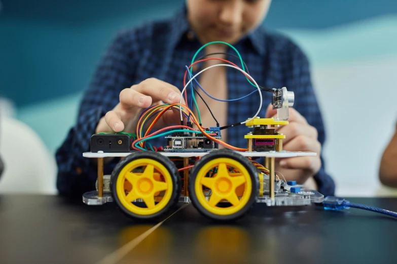 a person sitting at a table working on a robot, shutterstock, kinetic art, driving in a little toy car, arduino, 🦑 design, schools