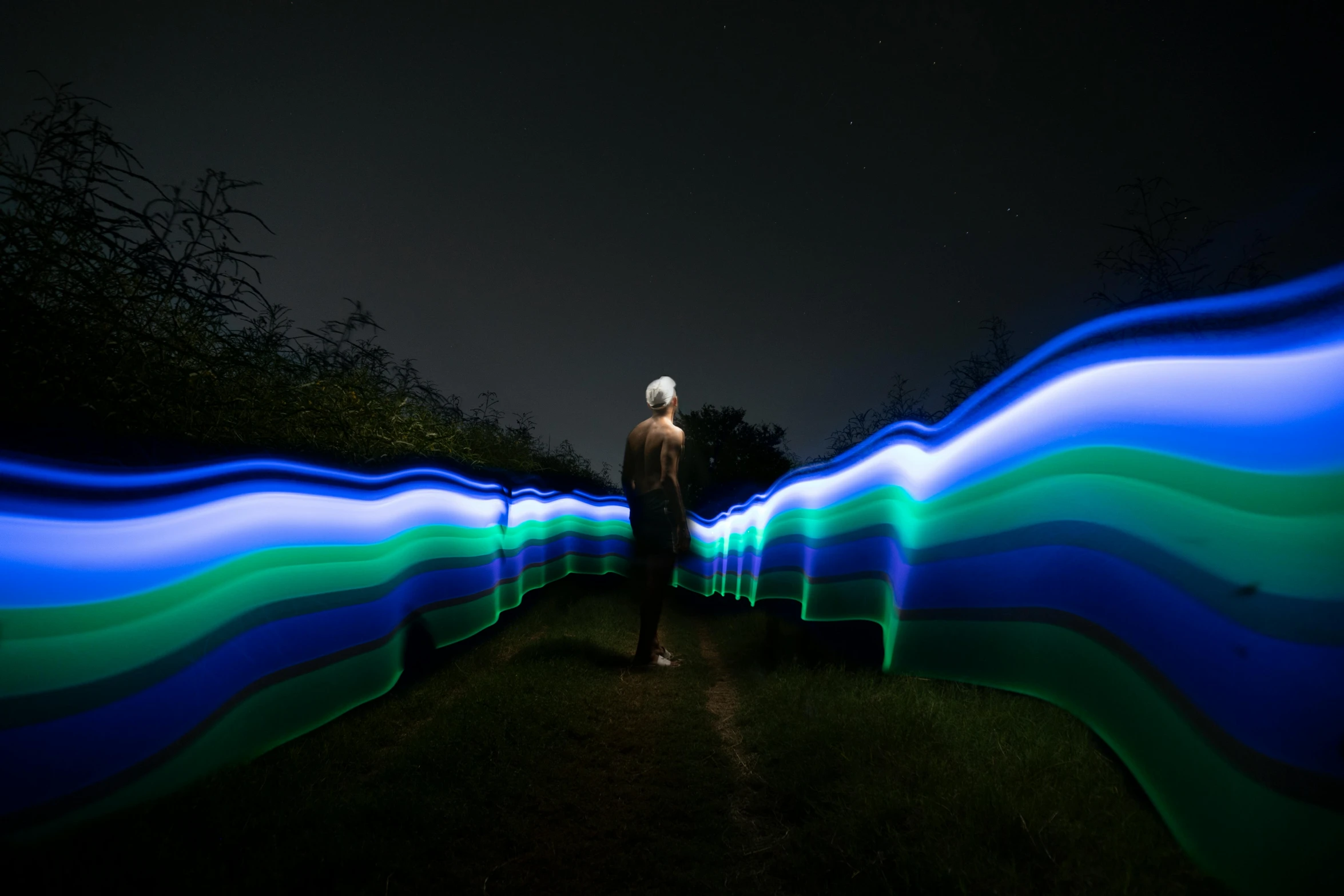a person standing in a field with blue and green lights, a picture, unsplash, interactive art, leading lines, flowing lines, in the woods at night, radiant slime trail