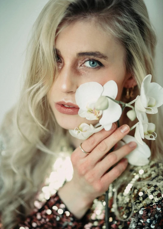 a woman holding a flower in front of her face, inspired by Elsa Bleda, white orchids, platinum blonde, non binary model, gilded. floral