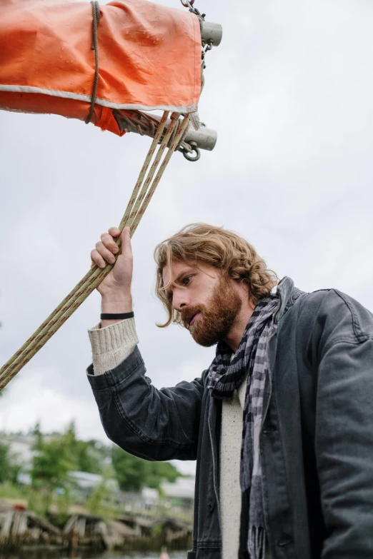 a man standing on top of a boat next to a body of water, by Jan Tengnagel, trending on unsplash, renaissance, braided beard redhead dreadlocks, hanging from a hot air balloon, ( ( theatrical ) ), wearing a scarf