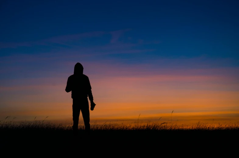 a person standing in a field at sunset, pexels contest winner, hooded figure, orange and blue sky, dark. no text, silhouette of a man