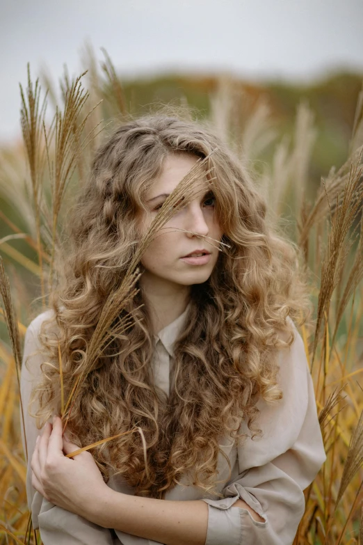 a woman sitting in a field of tall grass, inspired by Elsa Bleda, trending on pexels, renaissance, curly middle part haircut, long brown puffy curly hair, autumn season, like a catalog photograph