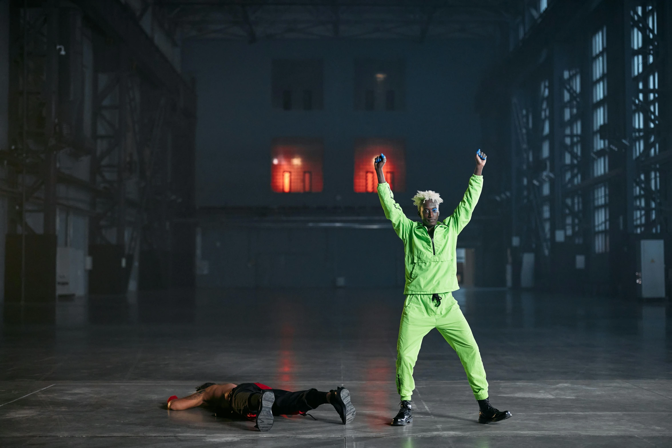 a man standing on top of a floor next to a person laying on the ground, inspired by Bruce Nauman, unsplash, video art, green rubber suit godzilla, in a dark warehouse, die antwoord style wear, performance