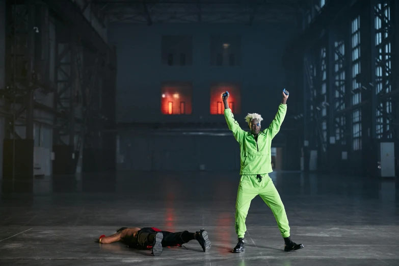 a man standing on top of a floor next to a person laying on the ground, inspired by Bruce Nauman, unsplash, video art, green rubber suit godzilla, in a dark warehouse, die antwoord style wear, performance