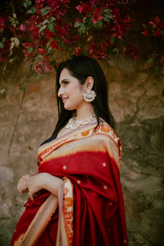 a woman in a red sari standing in front of a stone wall, an album cover, inspired by Raja Ravi Varma, pexels contest winner, silver jewellery, profile image, shawl, modeled