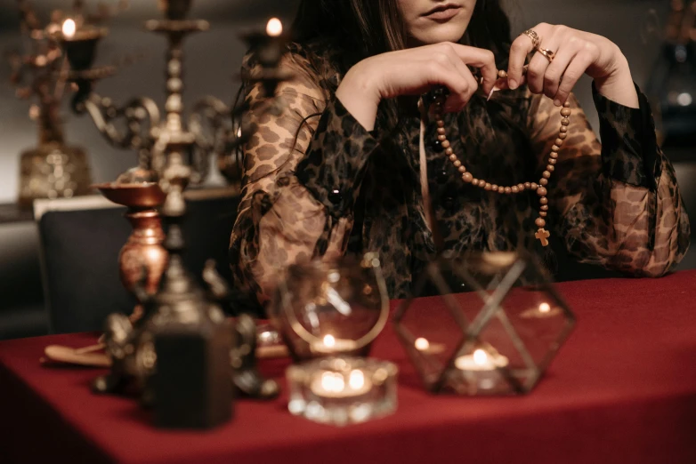 a woman sitting at a table with a red table cloth, inspired by Elsa Bleda, trending on pexels, renaissance, jewelry made of bones, black pearls and golden gems, ritual occult gathering, wearing brown robes