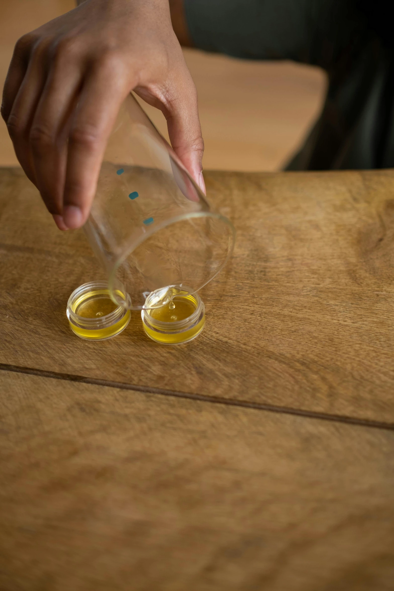 a person pouring something into a glass on top of a wooden table, made of wax and oil, healing pods, olive oil, oil on oak wood