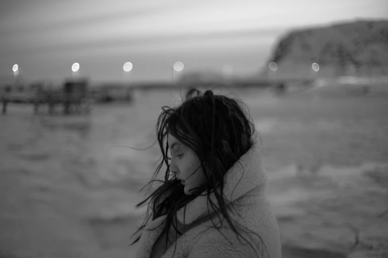 a woman standing in front of a body of water, a black and white photo, by Sergio Burzi, sza, cold freezing nights, windblown dark hair, on a beach