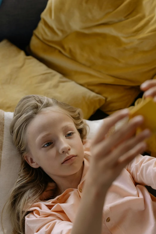 a little girl laying on a couch reading a book, a picture, pexels, checking her cell phone, yellow, movie still frame, square