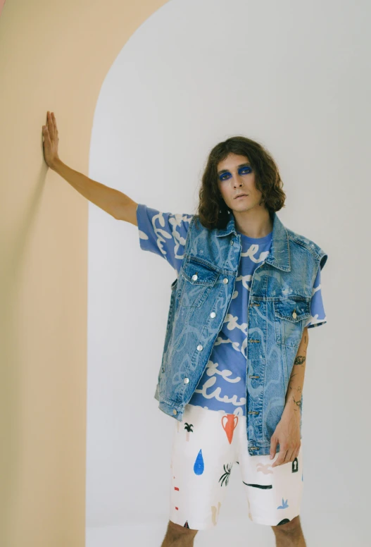 a man standing in front of a white wall, an album cover, by Olivia Peguero, graffiti, wearing double denim, an epic non - binary model, wearing a vest, patterned clothing