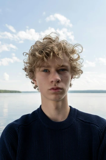 a young man standing in front of a body of water, an album cover, trending on pexels, photorealism, pale skin curly blond hair, finland, headshot profile picture, portrait of 14 years old boy