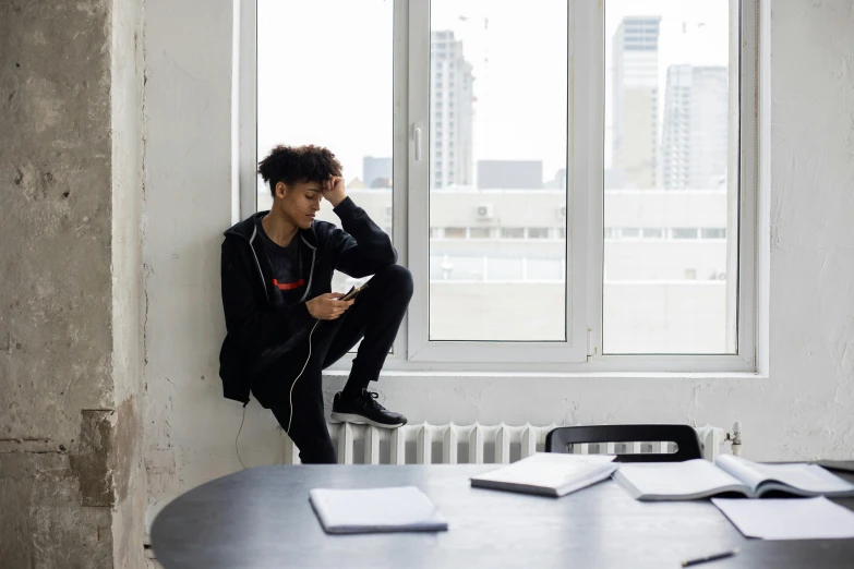 a man sitting on top of a table next to a window, trending on pexels, black teenage boy, phone background, androgynous person, bored