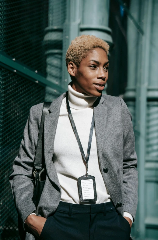 a close up of a person standing near a fence, grey turtleneck coat, short blonde afro, walking to work with a briefcase, wearing track and field suit