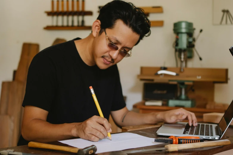 a man sitting at a table working on a laptop, a drawing, pexels contest winner, brian pulido, in a workshop, writing on a clipboard, carpenter