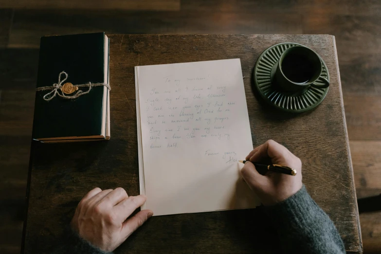 a person sitting at a table writing on a piece of paper, by Emma Andijewska, pexels contest winner, letterism, jaquet droz, cardboard, casey cooke, fan favorite