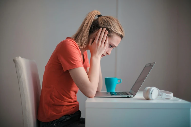 a woman sitting at a table with a laptop in front of her, pexels, devastated, wearing an orange t shirt, nursing, student